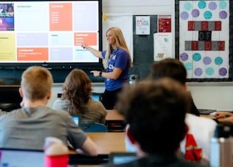 Person at front of classroom pointing to screen while teaching room of students.