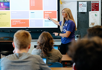 Teacher in purple shirt at front of classroom of students pointing to a screen.